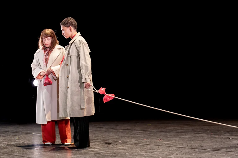 Zwei Schauspieler in Trenchcoats stehen auf einer Bühne, verbunden durch ein Seil mit roten Tüchern. Dunkler Hintergrund.