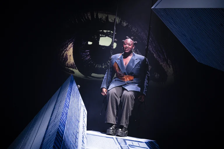 Ein Mann in einem modernen Theaterstück steht vor einem surrealen Hintergrund, der wie ein Auge aussieht. Dunkle Beleuchtung verstärkt die Stimmung.