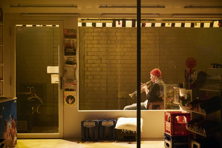 Ein Musiker spielt Flöte in einem Schaufenster eines Ladens. Die Szene ist warm beleuchtet, eine ruhige abendliche Atmosphäre.