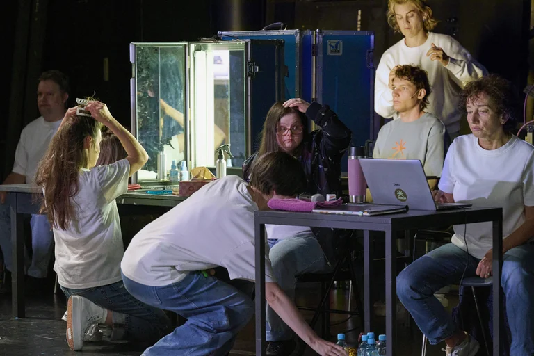 Eine Gruppe von Menschen probt auf einer Bühne. Sie sitzen um einen Tisch mit einem Laptop und Spiegel.